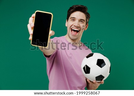 Young fun man 20s fan wear basic pink t-shirt cheer up support football sport team hold in hand mobile cell phone soccer ball watch tv live stream scream isolated on dark green color background studio