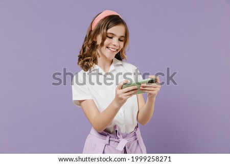 Little kid girl 12-13 year old in white shirt using play racing on mobile cell phone hold gadget smartphone for pc video games isolated on purple background studio Childhood children lifestyle concept