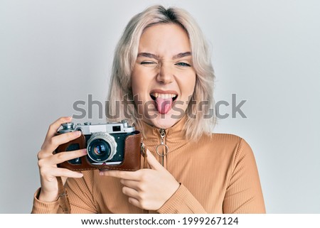 Young blonde girl holding vintage camera sticking tongue out happy with funny expression. 