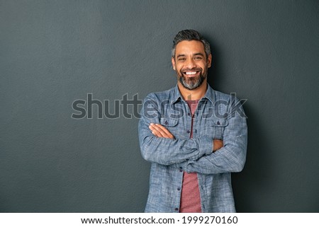 Handsome mid adult man with beard and crossed arms looking at camera. Mature middle eastern man isolated against grey wall and smiling. Satisfied indian guy looking at camera with a big laugh. Royalty-Free Stock Photo #1999270160