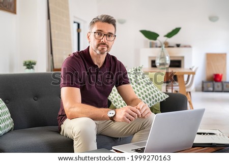 Portrait of mature man wearing spectacles and working from home. Mid adult man sitting on sofa while working on laptop and looking at camera. Businessman using laptop in telecommuting from home. Royalty-Free Stock Photo #1999270163