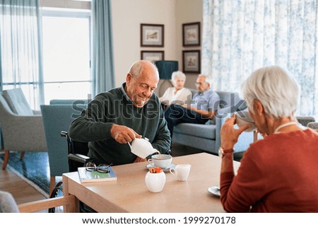 Smiling senior man pouring tea in cup from teapot in care facility with wife sitting at table in the common area. Elderly man in care centre sitting on wheelchair serving tea during a visit. Royalty-Free Stock Photo #1999270178
