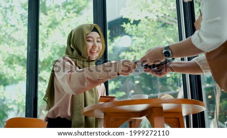 Young beautiful muslim woman wearing hijab sitting near glass window in coffee shop with bakery and coffee cup