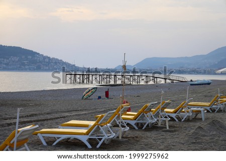 Empty sun loungers on a sandy beach against the backdrop of a pier, sea and mountains, evening, calm sea