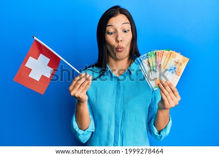 Young latin woman holding switzerland flag and franc banknotes making fish face with mouth and squinting eyes, crazy and comical. 