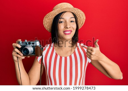 Young latin woman wearing summer hat holding vintage camera smiling happy and positive, thumb up doing excellent and approval sign 