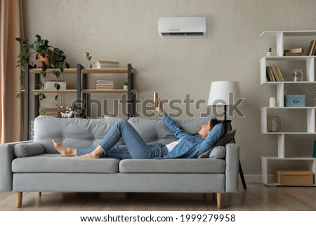 Full length relaxed young happy woman homeowner lying on comfortable sofa turning on air conditioner with remote controller, breathing fresh air, enjoying lazy weekend time, meditating in living room. Royalty-Free Stock Photo #1999279958