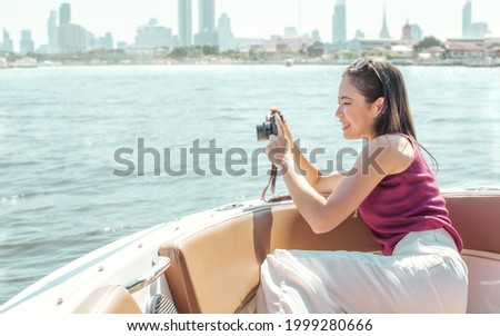 Beautiful long hair Asian woman wearing casual clothes with sunglasses, holding camera take landscape sea scenery photo while sitting on boat, smiling with happiness during summer outdoor vacation