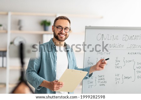 Portrait of happy young male teacher standing near blackboard with English grammar rules, conducting internet lesson. Positive tutor giving oreign language class on video call or web conference Royalty-Free Stock Photo #1999282598