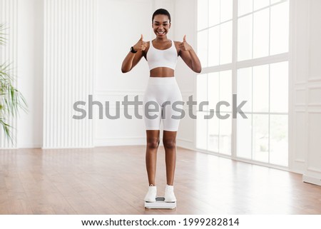 Excited black woman standing on scales at home or fitness studio, happy with result of her diet and exercises, showing thumbs up, free copy space. Emotional lady achieving weight loss goal, success Royalty-Free Stock Photo #1999282814