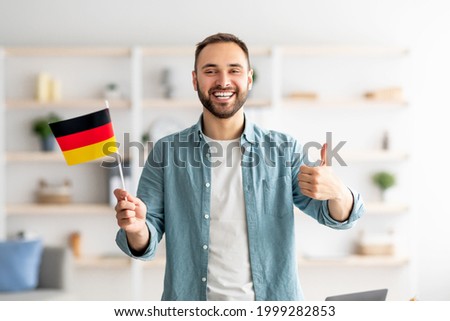 Happy Caucasian man showing thumb up and flag of Germany, posing and smiling at camera indoors. Cheerful millennial student recommending foreign education, learning German language Royalty-Free Stock Photo #1999282853