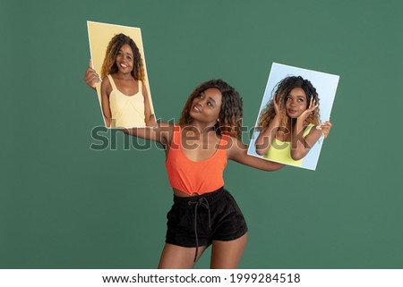 Young happy african girl holding her portraits, pictures isolated on green background. Smiling or thoughtful. Concept of human emotion, facial expression.