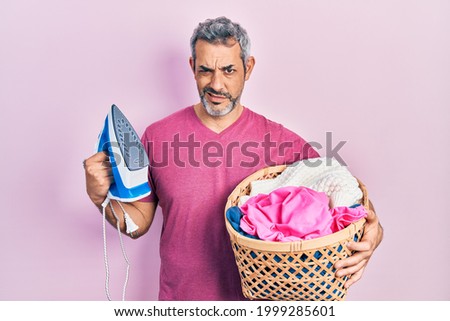 Handsome middle age man with grey hair holding electric steam iron holding laundry basket clueless and confused expression. doubt concept. 