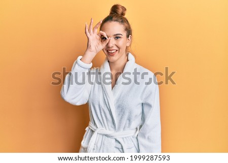 Young blonde woman wearing bathrobe doing ok gesture with hand smiling, eye looking through fingers with happy face. 