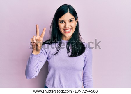 Young hispanic woman wearing casual clothes smiling looking to the camera showing fingers doing victory sign. number two.  Royalty-Free Stock Photo #1999294688