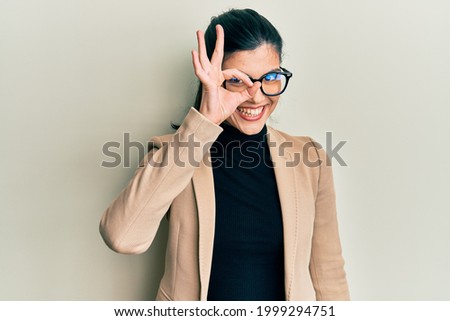 Young hispanic woman wearing business style and glasses smiling happy doing ok sign with hand on eye looking through fingers 