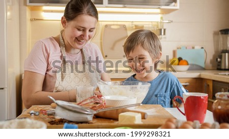 Happy laughing boy with mother playing and fooling around while making dough for biscuits at home. Children cooking with parents, little chef, family having time together, domestic kitchen.