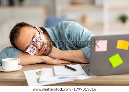 Lazy unproductive young guy wearing funny sticky notes with open eyes on his glasses, sleeping at workplace. Tired overworking millennial man falling asleep at home office Royalty-Free Stock Photo #1999313816