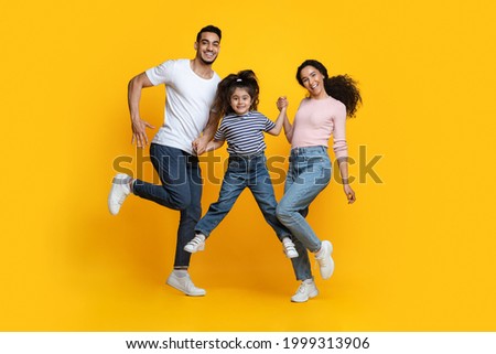 Portrait Of Cheerful Excited Middle Eastern Family Of Three Holding Hands While Jumping In Air Over Yellow Background In Studio, Happy Arab Parents And Little Daughter Having Fun Together, Copy Space