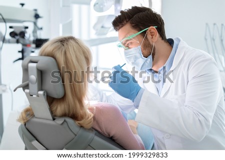 Man dentist in face mask and glasses doing treatment for patient blonde lady, holding dental tools, wearing rubber gloves. Stomatology, dentistry, modern dental clinic concept Royalty-Free Stock Photo #1999329833