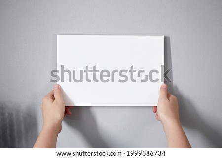 White canvas in female hands with gray wall background. Woman hanging blank picture mockup on wall. Wall decor