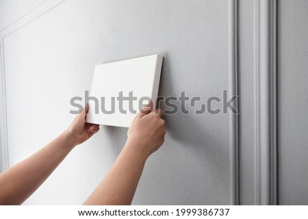White canvas in female hands with gray wall background. Woman hanging blank picture mockup on wall. Wall decor
