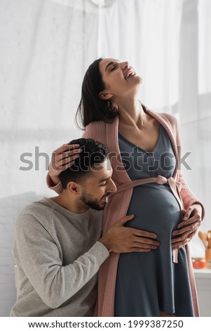 happy young man touching belly of pregnant woman in kitchen