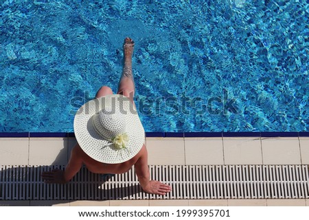Woman in a sun hat sits on the edge of the pool and swings her legs, top view. Summer holidays, tanning and vacation at a tourist resort