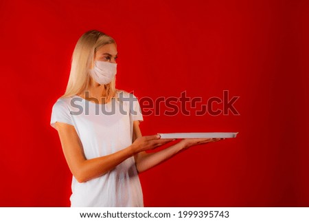Blondy nurse girl in face mask presenting on tray looks aside. Copy space photo of slim blonde in white medical mask and white outfit on matte red background holding mockup plate