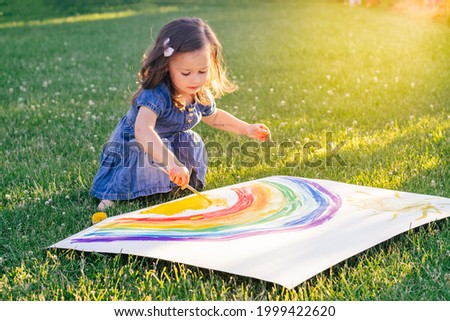 Little girl 2-4 years old paints rainbow and sun on large sheet of paper sitting on green lawn in sunlight