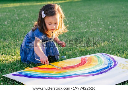 Little girl 2-4 years old paints rainbow and sun on large sheet of paper sitting on green lawn