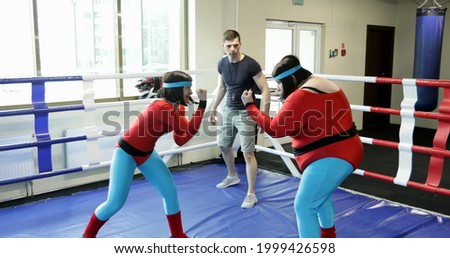 Fat and thin women train in the boxing ring with a trainer. A thin athlete runs away from a boxing rival.