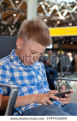 Little boy sitting in airport departure hall and waiting for his flight. Kid at airport is playing tmobile phone. Child in waiting room before vacation.