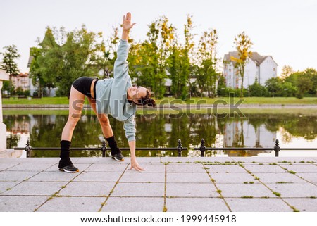 Young sportswoman do complex of stretch exercise before running in a park near the lake in the early summer morning