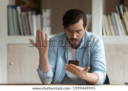 Angry nervous young 30s male user in eyeglasses looking at smartphone screen, feeling dissatisfied with malware spam scam message, bad device work, lost data, getting awful news in modern office. Royalty-Free Stock Photo #1999449995