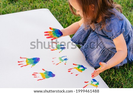 Little girl 2-4 draws leaving rainbow colors child's handprints on large white sheet of paper
