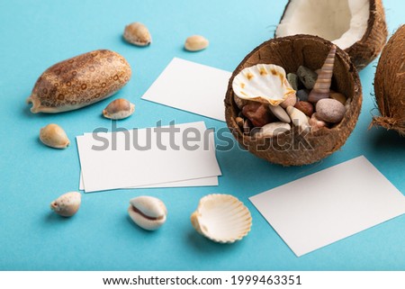 White paper business card with coconut and seashells on blue pastel background. Side view, copy space. Tropical, healthy food, vacation, holidays concept.