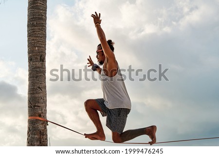 young latin man balancing on the slackline kneeling on the slackline on two palm trees in the middle of the beach on a sunny day, outdoor sport concept. Royalty-Free Stock Photo #1999476245