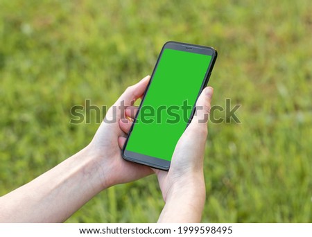 Mobile phone mock up with green screen template in female hand over green lawn. Smartphone with empty display.