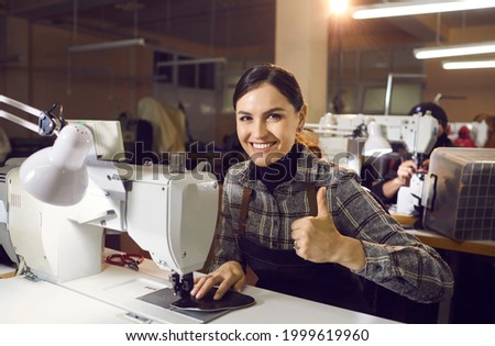 Portrait of happy young woman sitting at shoe sewing machine, smiling, looking at camera, giving thumbs up. Footwear factory worker satisfied with good salary, material quality and working conditions