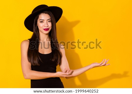 Photo of happy young nice woman hold hands empty space wear hat smile isolated on yellow color background