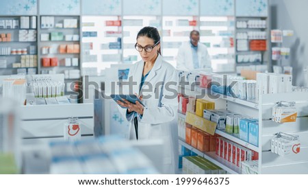 Pharmacy Drugstore: Beautiful Caucasian Pharmacist Uses Digital Tablet Computer, Checks Inventory of Medicine, Drugs, Vitamins, Health Care Products on a Shelf. Professional Pharmacist in Pharma Store Royalty-Free Stock Photo #1999646375