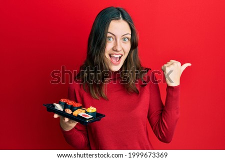 Young brunette woman showing a plate of sushi pointing thumb up to the side smiling happy with open mouth 