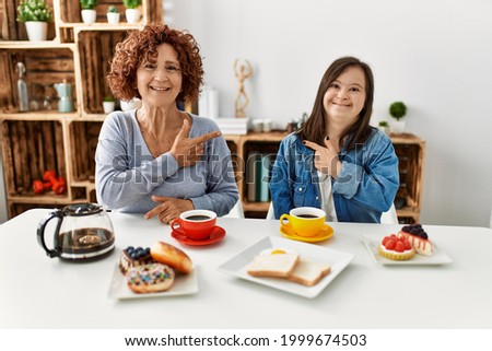 Family of mother and down syndrome daughter sitting at home eating breakfast cheerful with a smile of face pointing with hand and finger up to the side with happy and natural expression on face 