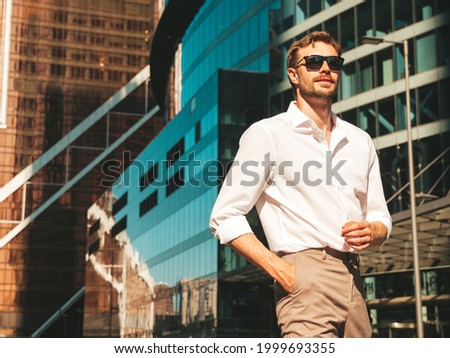 Portrait of handsome confident stylish hipster lambersexual model.Modern man dressed in white shirt. Fashion male posing in the street background near skyscrapers in sunglasses. Outdoors at sunset  Royalty-Free Stock Photo #1999693355