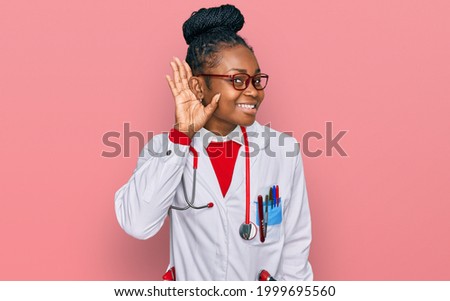 Young african american woman wearing doctor uniform and stethoscope smiling with hand over ear listening an hearing to rumor or gossip. deafness concept. 