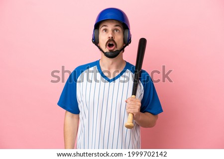 Young caucasian man playing baseball isolated on pink background looking up and with surprised expression
