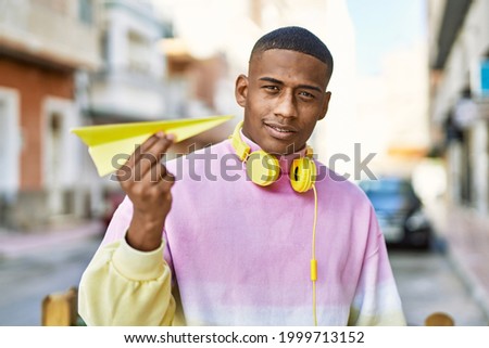 Young african american man using headphones holding paper airplane at the city.