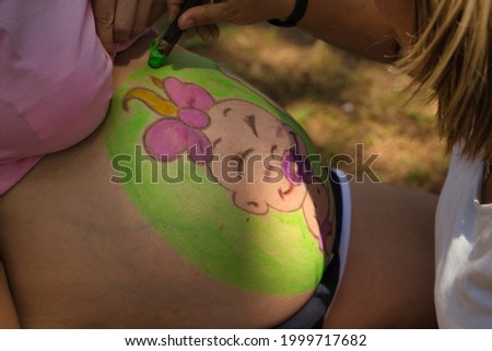Person painting the belly of a pregnant woman during the celebration of a pregnancy party. Outdoors on a sunny day.