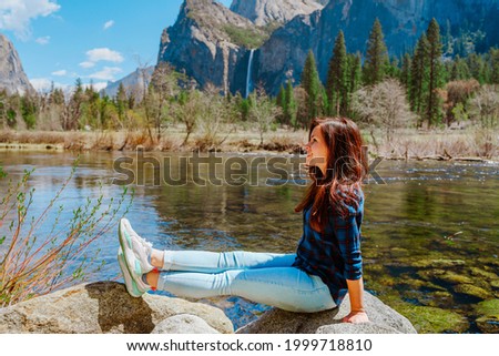 A beautiful young woman in a blue shirt is resting by the river in the mountains in Yosemite National Park
